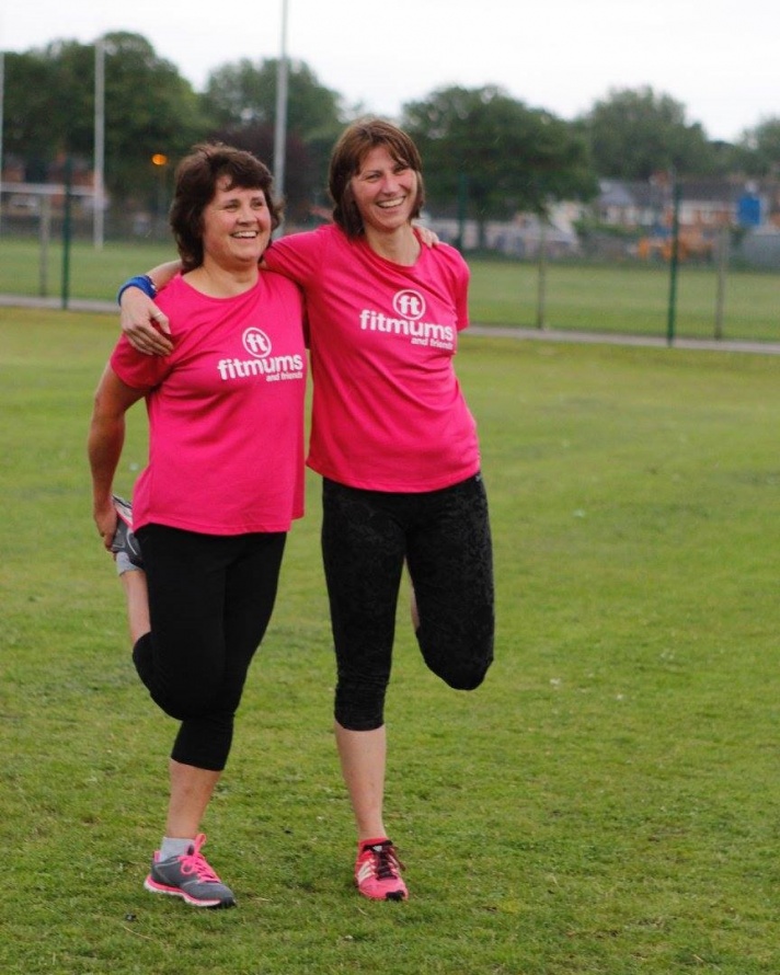Two laughing women in pink Fitmums t-shirts hold each other up to stretch their legs